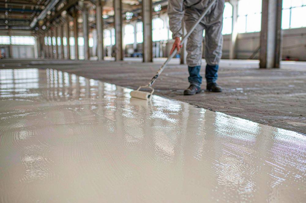 worker applying solvent-free high-solids epoxy floor coating with a long-handled roller on a concrete floor